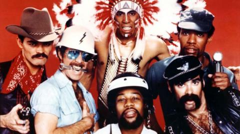 The Village People pose in a group studio shot wearing their various costumes against a red background, taken in 1977.