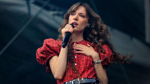 Holly Humberstone performing at a festival in August. Holly is a 22-year-old woman with long curly brown hair which she wears loose. She has blue eyes and holds a hand to her chest as she sings into a microphone. She's styled in a red tartan-look dress with puff sleeves and a studded black leather belt. 