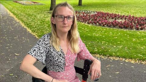 Laura Earl is sitting in a wheelchair and looking at the camera with a neutral expression on her face. She is in a park, on the grey tarmac path, with a grassy area and red flower bed behind her. She has long blonde hair, glasses and is wearing a pink flowery dress.