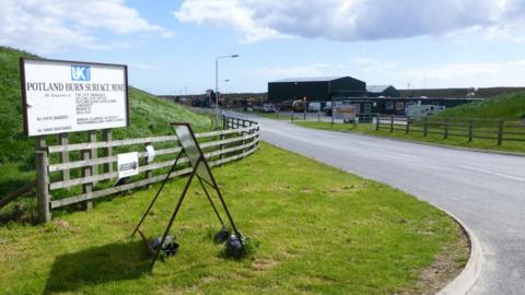 The entrance to Potland Burn Surface Mine. There is wooden fencing and grassy verges either side of a road leading into the site. There is a white sign to the left saying Potland Burn Surface Mine.