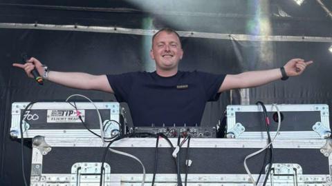 A man wearing a black t-shirt standing behind DJ decks. He has his arms outstretched to the side with his fingers pointing outwards.