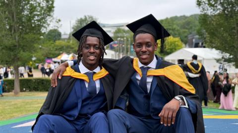 Two young men in Navy blue suits wearing black and yellow robes and black mortarboards