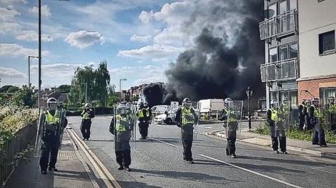 Police officers in riot gear spread out across a road with black smoke visible in the background. 