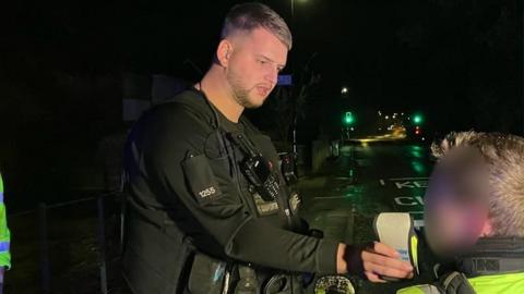 A traffic officer dressed in black uniform holds out a breathalyser device while a young man, whose face is blurred, blows into it on a dark street.