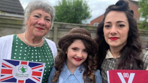 An older woman, a girl and a younger woman stand together smiling. They all have their hair and make-up done in a 1940s style and are also wearing retro dresses. They are holding VE Day and Union Jack flags. 
