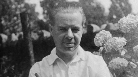 Clement Ernest Kille is seen in black and white wearing a white shirt and standing in front of a flower bush. He has his hair combed back and is smiling.