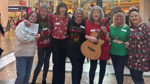 Seven women stand in the middle of a shopping centre, they are all wearing festive jumpers. The woman in the middle holds a guitar