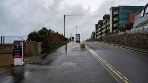 A digital screen which appears to be the size of a telephone box can be seen on the pavement at the side of the beach, with apartments seen on the other side of the road.