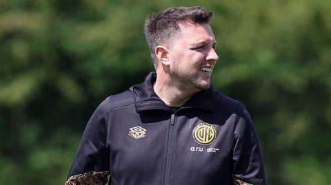 A smiling Mark Bonner at the club's training ground wearing a black tracksuit top with the club badge on