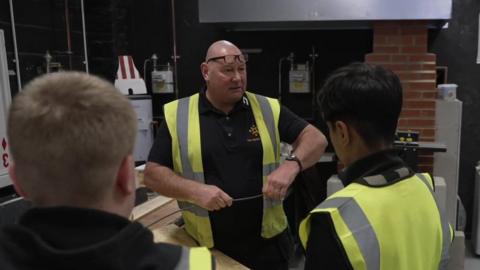 A man wearing a high visibility jacket is talking to a group of young boys, also wearing high visibility jackets. They appear to be in an industrial facility.