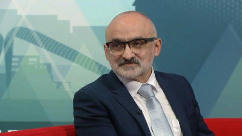 A man with a bald head, a white beard and dark-framed glasses in a blue suit jacket, white shirt and pale blue tie sat on a red sofa with a blue background