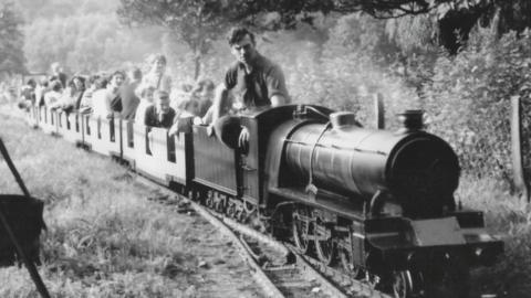 A black and white image of a miniature railway with at least seven carriages filled with parents and children and a train driver in rolled up shirt sleeves driving the miniature engine.  In the background can be seen grass and trees.