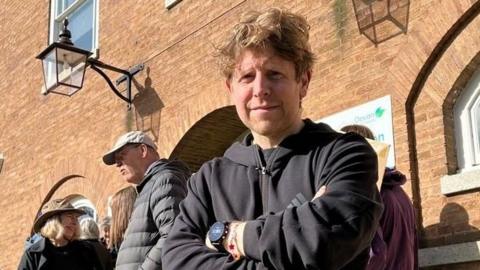Josh Widdicombe on steps outside County hall with other protesters behind. He is wearing a black hoodie and a black watch and has his arms folded.