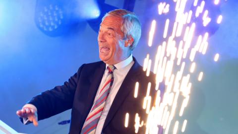 Reform leader Nigel Farage wearing a dark blue suit jacket, white shirt and stripy tie while smiling. A gold firework is let off in front of him