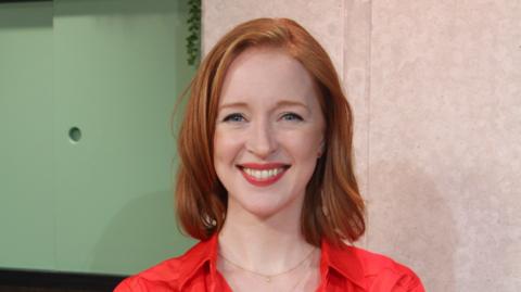 Finance expert Iona Bain in the Morning Live studio, wearing a red top and standing in front of a green and beige wall.