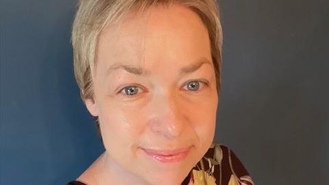 Dr Helen Evans-Howells takes a selfie. She is standing against a dark blue wall, has short blonde hair, blue eyes and smiles at the camera. She is a black blouse with a floral print.