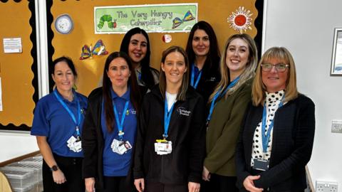 Seven women standing in a nursery setting smiling. There are boards behind them with 'The Very Hungry Caterpillar' pictures and writing on. They all have blue lanyards around their necks and most of them are wearing either blue polo shirts or black fleece's with the centre's branding on it.