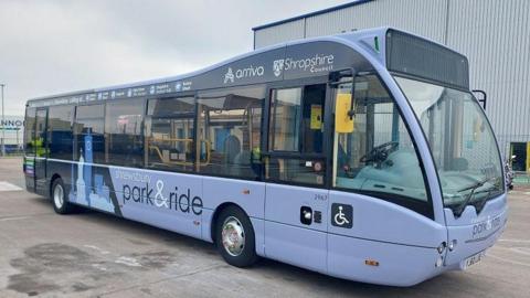 A lilac coloured bus with Shropshire Council and Arriva logos above the windows. There is a sign on the side that says "Shrewsbury park and ride" and there is a decal that shows silhouettes of some of the town's buildings.