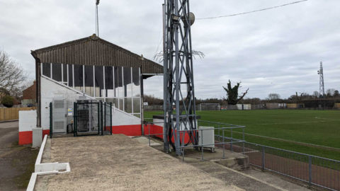 Part of the ground at Stag Meadow, with a covered terrace to the left of the picture and the grass pitch to the right, with the bottom part of a floodlight in the centre of the picture.