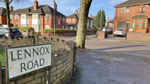 The corner of Lennox Road with a road sign showing the street name. The residential road has houses, parked cars and tree trunks, with wide pavements on both sides.