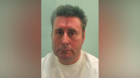 A mugshot image of a middle-aged male prisoner wearing a white top.