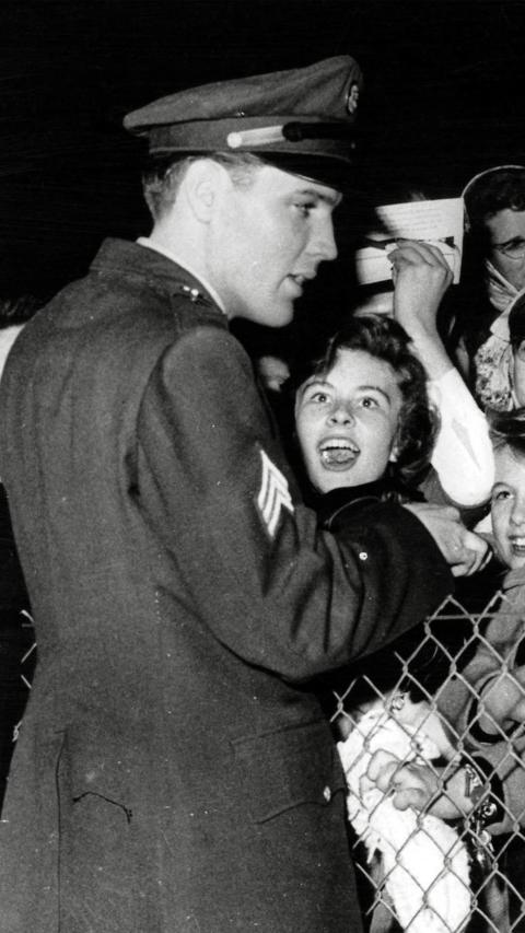 A black and white photo of Elvis on one side of a chain-link fence, with excited fans on the other.