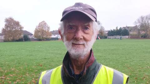 A man with a white beard and hair, wearing a cap and a yellow high-vis jacket