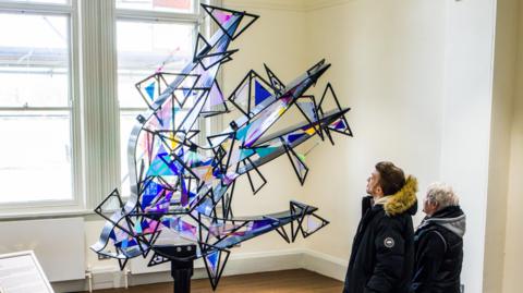 Two people are looking up at the artwork. Shards of blue and purple glass are arranged in a metal frame that resembles a swallow in flight. It is placed in a white room, in front of a window with sunlight streaming through, which means the colours of the glass are illuminated.