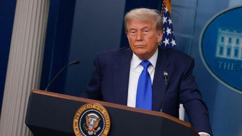 Donald Trump wears a dark suit and blue tie as he stands at a podium in the White House briefing room.