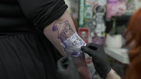 The image shows the hand of a tattoo artist wearing black gloves, peeling off the stencil of a tattoo of a woman with a hat. The arm of the person receiving the tattoo is partially covered by a black top that looks to be a black dress.