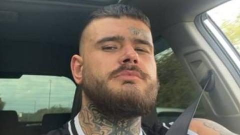 A selfie of a man sitting in a car. The trees in the background are blurred showing the car is moving. He is wearing a seatbelt and looking into the lens. He has a bear, a neck tattoo and a small tattoo above his eyebrow.