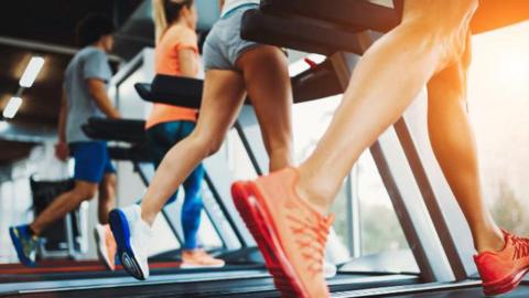 Close up shot of people running on treadmills. The person closest to the camera is wearing bright orange trainers. There is a large window in front of the equipment.