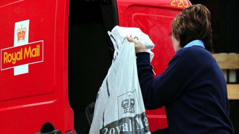 With her back to the camera a shorthaired woman wearing a navy sweater lifts a grey sack into the back of red Royal Mail van. 