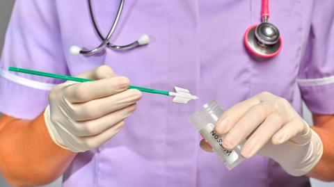 A nurse wearing a purple nurse's outfit is holding a plastic tube and a cervical paintbrush. She has white rubber gloves on.