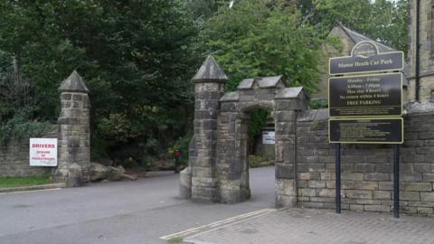 A grand entrance to a park with a sign to the right that reads: Manor Heath Car Park. There are trees and stone buildings and a road leading between stone pillars.
