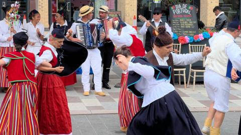 Group of performers in colorful traditional dress dancing and playing music, including an accordion and guitar, at a street event with bunting and a sign reading “Dia de Portugal – Presents.”