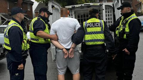 The back of a tall man, wearing a white short sleeved T-shirt and grey tracksuit shorts. He is also wearing handcuffs and being escorted to a police van by four officers. There are two male officers on the man's left wearing all black and yellow hi-vis vests and black caps, and on the man's right is a female officer and male officer wearing the same