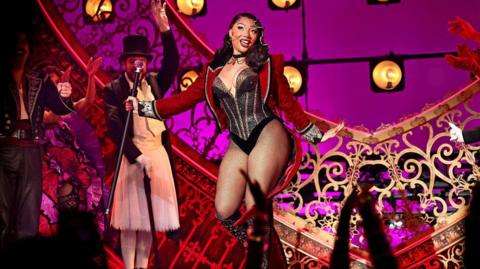 Megan is wearing a red cropped jacket, a black sparkly leotard with silver stripes. She is wearing fishnet tights and black knee high boots. She is holding a black cane and smiling a the crowd. She is on stage, behind her is gold and red decorations. To her left is other members of the cast, a man in a suit and top had and a man in all black. 