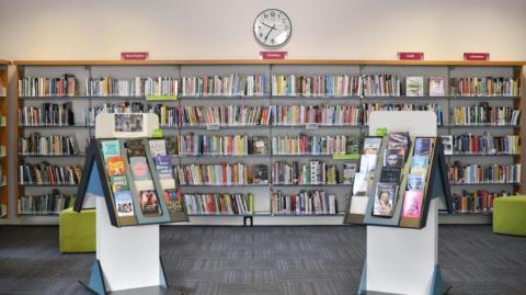 The inside of a library. There are two small book cases in the foreground, more like a sales stand in that they are triangular and allow books to be shown with the covers facing out. A large wall-to-wall shelf unit stands behind them, all full of books and with red signs above denoting subject areas. There are two green pouffes in the room, which has white walls and grey patterned carpet.
