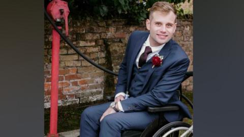 Liam in his wheelchair wearing a wedding style petrol blue suit - with a red rose on the lapel. He looks at the camera and smiles. He is on the pavement next to a brick wall.
