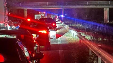 A queue of traffic on the motorway, it is dark and there are lots of red brake lights. There is a blue light in the distance. 