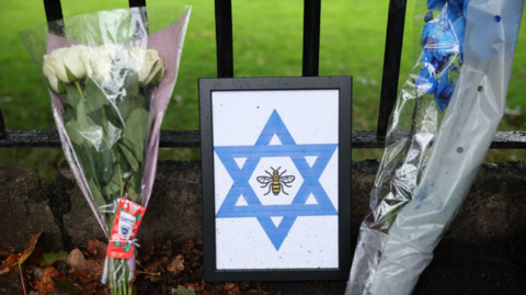 Floral tributes and the Star of David are displayed at the attack site. The tributes have been placed on the ground and are resting next to a black metal fence. Green grass can be seen behind the gate.