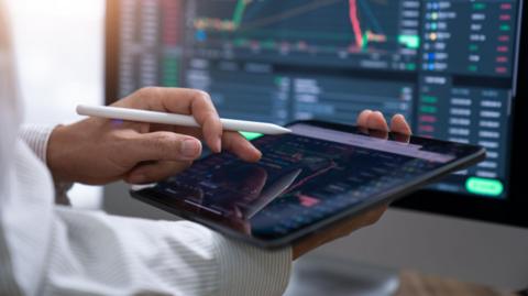 A person reviews financial information in a tablet, with a computer screen mirroring the data.