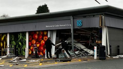 A Co-op shop that has the frontage destroyed leaving bits of debris strewn across the floor. A telehandler can be seen in front of it with it's two prongs in the air and a sign is in front of that with "Co-op" on it and its opening times.