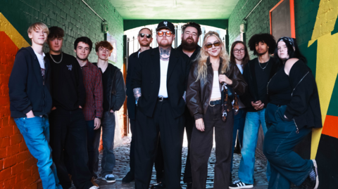 The band the Lottery Winners stand in an alley with dark green coloured walls while flanked by two groups of young people. They are posing for a promotional photograph. 