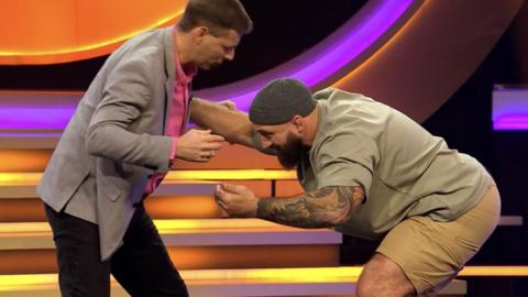 Blankety Blank contestant, Glen gets ready to form a scrum with former rugby start Joe Marler. They are in a TV studio.
