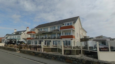 The outside of the Cobo Bay Hotel in Guernsey. It's a three story building with balconies and lots of outdoor seating. There is also a sign with the company logo at the front entrance. 