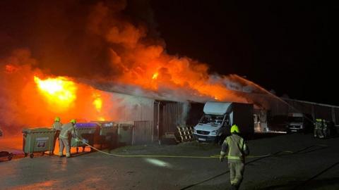 Firefighters along the side of a large corrogated building - they are spraying water onto flames seen filling the inside of the building.