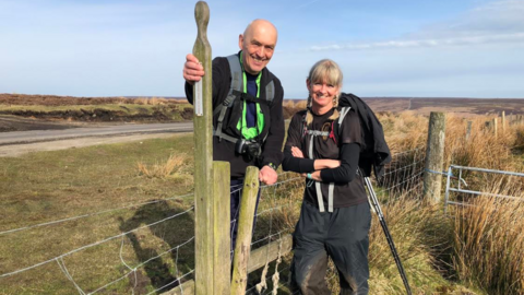 Two walkers, wearing walking gear stand by a wooden stile in the North York Moors. In the background, hills covered in heather can be seen.