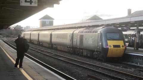 A train at a railway station. Railway tracks run through the train station. A man is standing on the platform taking a picture of the train. 